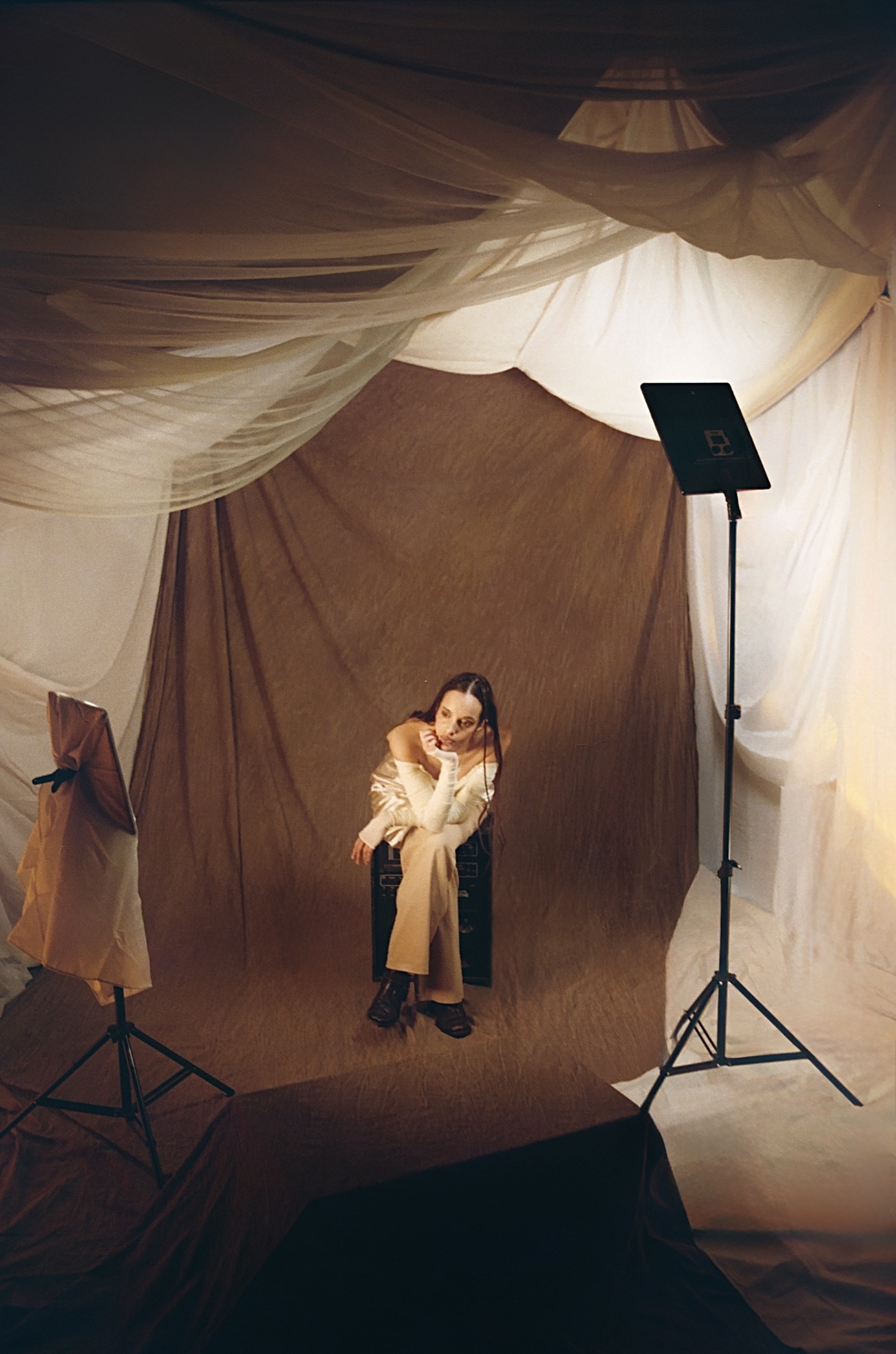 Photographie de Constance Guivarc'h représentant Laure Sanchez, bassiste et chanteuse bordelaise, assise sur un tabouret dans un décor de shooting photo, habillé de draps beige et marron. Photographie de promotion pour son titre "Aux Absents" et sa tournée en première partie de Vanessa Paradis.