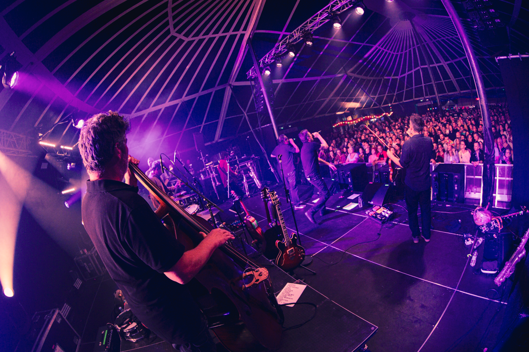 Le groupe de rock caravane français, Les Hurlements d'Léo, sur scène devant la foule. La photographie est prise depuis le côté jardin de la scène, avec un effet grand angle, et une colorimétrie violet-bleu.